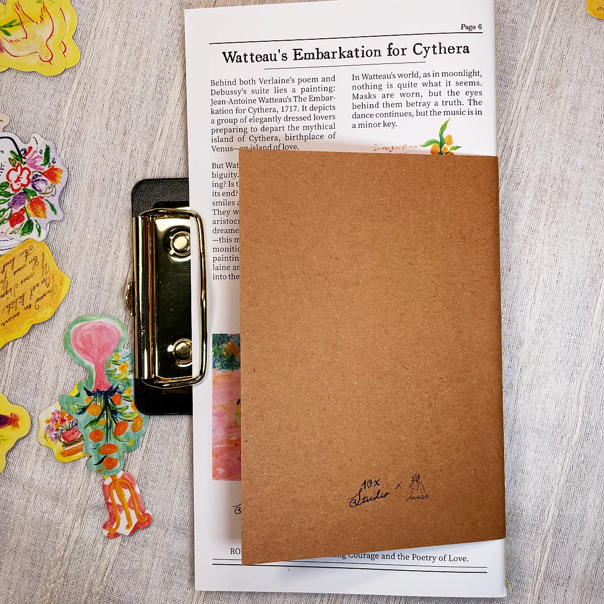Flat lay of the brown kraft paper notebook back cover with studio branding, clipped to a vintage newspaper page featuring art history text and surrounded by aesthetic stickers.