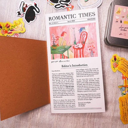 A printed card styled like a vintage 'Romantic Times' magazine page with a pink watercolor illustration of a chair and table, placed on top of a brown envelope. The card is surrounded by decorative stickers and a small tin on a light wood surface.