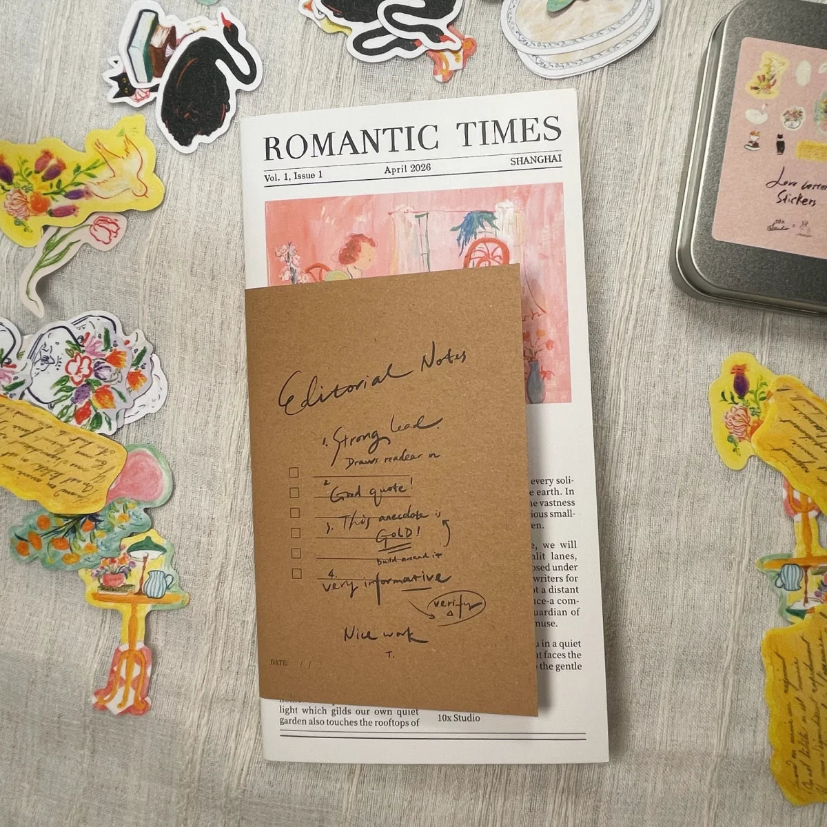 A kraft brown paper note pad with handwritten notes on top of a printed 'Romantic Times' magazine page, styled on a light wood surface surrounded by decorative stickers and a small tin.