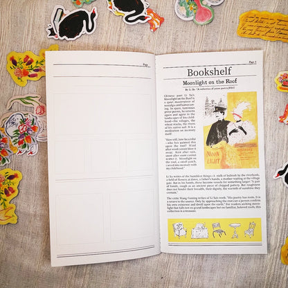 An open book or journal laid flat on a light wood surface, surrounded by colorful decorative stickers and paper cutouts. The left page has blank lined writing areas, and the right page shows a printed article titled 'Bookshelf' with text and a yellow illustration with a man and a woman.