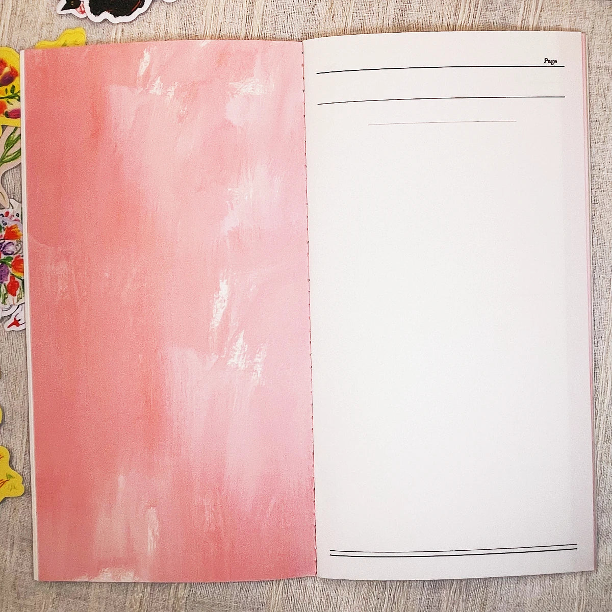 An open notebook or journal showing a textured pink left page and a blank white right page with thin black lines and the word 'Page' at the top right, photographed from above on a light wood surface.