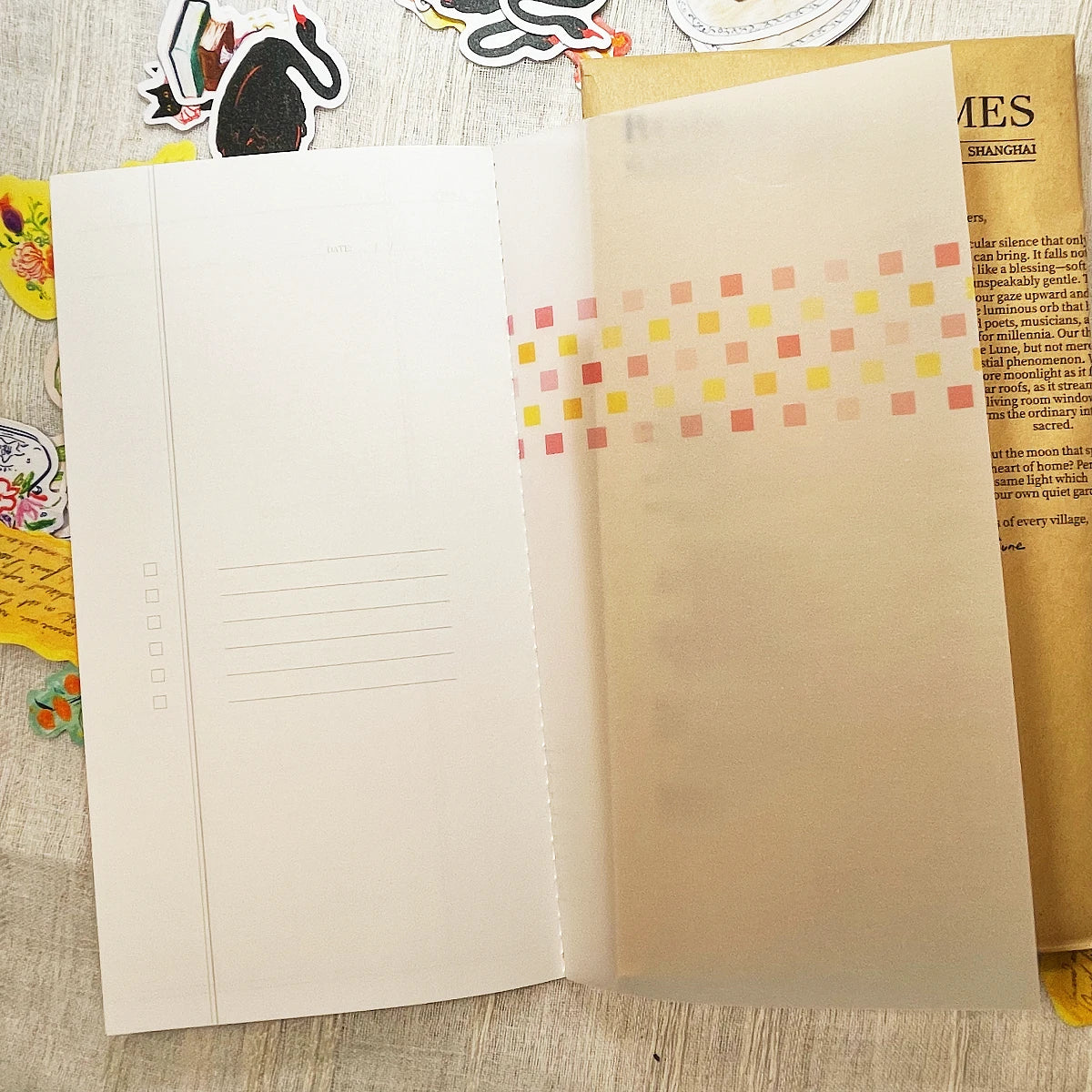 An open planner or notebook with blank lined pages on the left and a translucent beige right page decorated with a row of small pastel square stickers in pink, yellow, and orange. The notebook is photographed from above on a light wood surface with a few colorful stickers and papers partially visible around it.