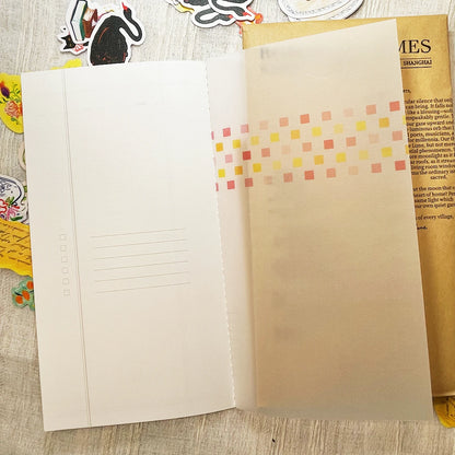 An open planner or notebook with blank lined pages on the left and a translucent beige right page decorated with a row of small pastel square stickers in pink, yellow, and orange. The notebook is photographed from above on a light wood surface with a few colorful stickers and papers partially visible around it.