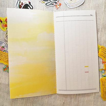 An open notebook or planner spread on a light wood surface, with a yellow watercolor wash on the left page and a lined grid page on the right. Decorative stickers are partially visible around the edges.